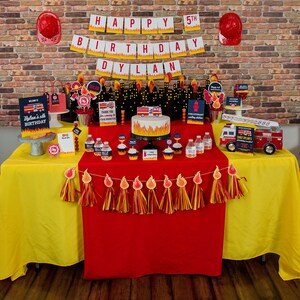 May include: A red and yellow fire truck themed birthday party table setting with a red tablecloth, yellow tablecloths, and a fire truck cake. The table is decorated with fire truck themed decorations, including a fire truck cake, fire truck cupcakes, and a fire truck banner. The table is also decorated with red and yellow streamers.