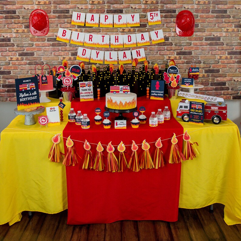 May include: A red and yellow fire truck themed birthday party table setting with a red tablecloth, yellow tablecloths, and a fire truck cake. The table is decorated with fire truck themed decorations, including a fire truck cake, fire truck cupcakes, and a fire truck banner. The table is also decorated with red and yellow streamers.