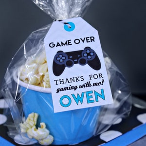 May include: A blue and white favor bag with a white tag that says "Game Over" with a black video game controller graphic. The tag also says "Thanks for gaming with me! Owen".