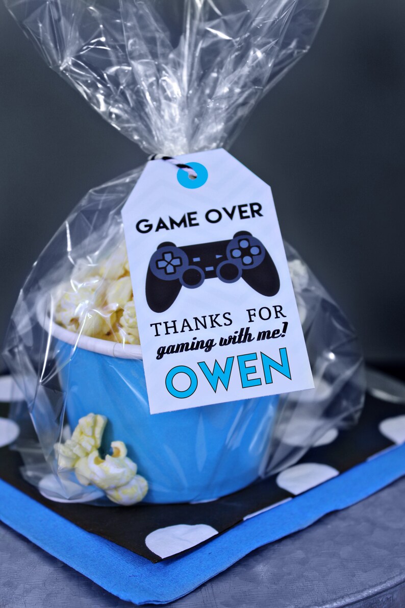 May include: A blue and white favor bag with a white tag that says "Game Over" with a black video game controller graphic. The tag also says "Thanks for gaming with me! Owen".