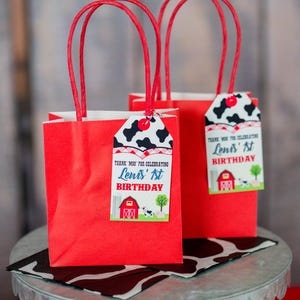 May include: Two red gift bags with white handles and cow print gift tags. The tags say "Thank you for celebrating  Leni's 1st Birthday".