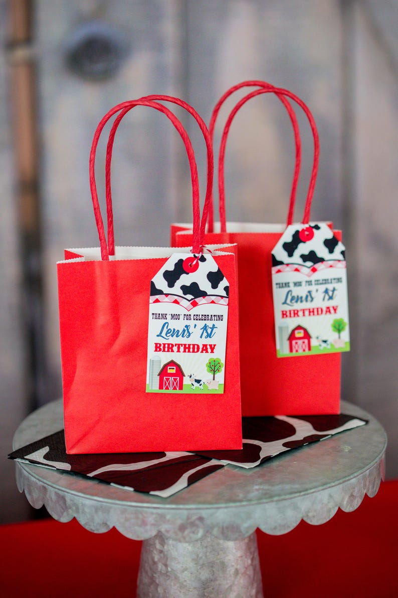 May include: Two red gift bags with white handles and cow print gift tags. The tags say "Thank you for celebrating  Leni's 1st Birthday".