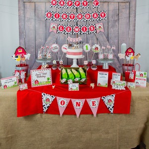May include: A red and white tablecloth with a banner that says "ONE" in black and white letters. The table is decorated with a farm-themed birthday party decorations, including a cake, cupcakes, and a banner that says "Happy 1st Birthday Lewis".