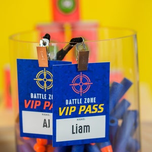 May include: Two blue and yellow VIP pass cards with a target symbol and the text "Battle Zone VIP Pass". The cards are hanging from clips and are in front of a clear container filled with blue and orange foam darts.