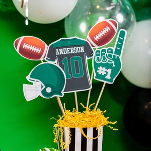 May include: A football-themed centrepiece with a green and black jersey featuring the name "Anderson" and the number "10". There is also a football, a football helmet, and a foam hand with the number "#1" on it. The centrepiece is in a black and white striped container with yellow shredded paper inside.
