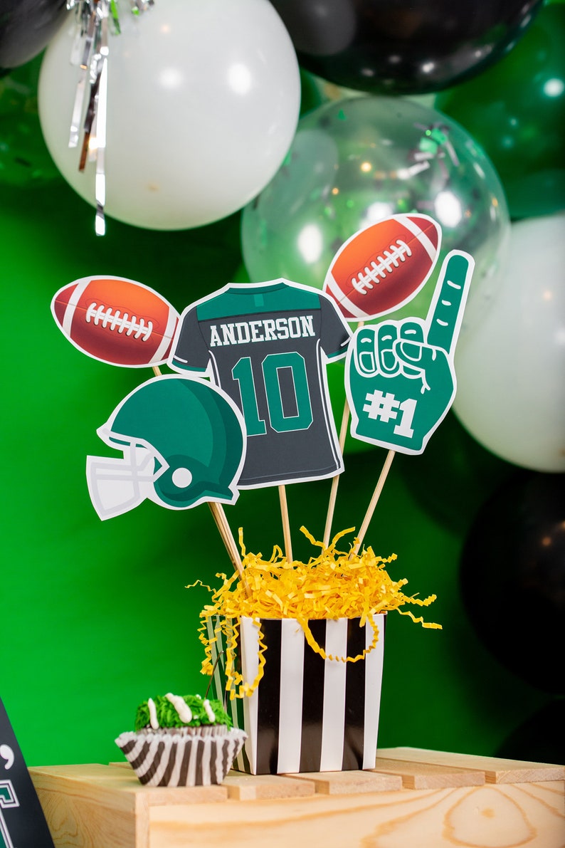 May include: A football-themed centrepiece with a green and black jersey featuring the name "Anderson" and the number "10". There is also a football, a football helmet, and a foam hand with the number "#1" on it. The centrepiece is in a black and white striped container with yellow shredded paper inside.