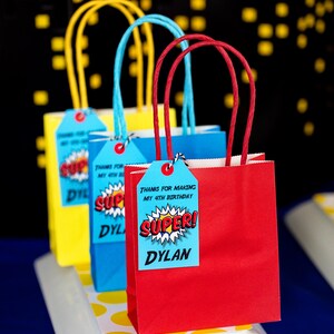 May include: Three red, blue, and yellow paper gift bags with handles. Each bag has a tag attached with the text "Thanks for making my 4th birthday SUPER! Dylan".