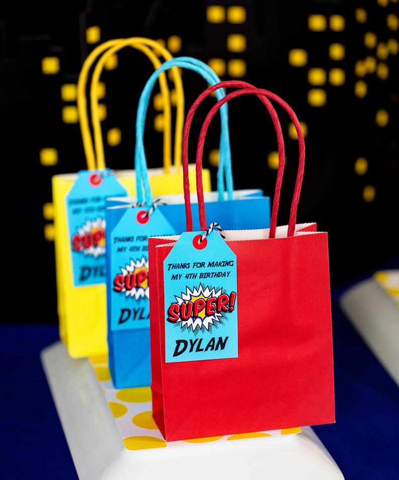 May include: Three red, blue, and yellow paper gift bags with handles. Each bag has a tag attached with the text "Thanks for making my 4th birthday SUPER! Dylan".