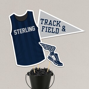 Könnte beinhalten: Marineblaue Leichtathletik-Dekorationen. Enthält ein Trikot mit dem Text "STERLING", eine Wimpel mit dem Text "TRACK FIELD &" und einen Laufschuh mit Flügeln. Die Dekorationen befinden sich in einem schwarzen Eimer.