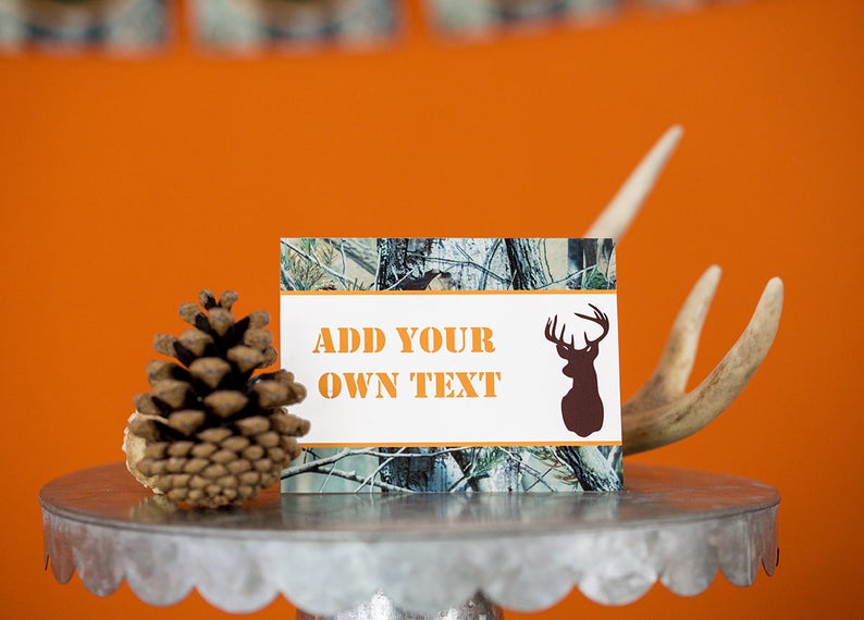 May include: A white card with a camouflage pattern and a brown deer silhouette. The card has the text "ADD YOUR OWN TEXT" in orange letters. The card is sitting on a silver cake stand with a pine cone and antlers.