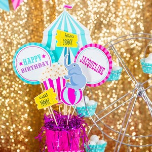 May include: A pink and blue circus-themed birthday party centerpiece with a tent, elephant, popcorn, and tickets. The centerpiece is in a pink bucket with pink and purple tinsel. The text "Happy Birthday" and "Jacqueline" are on the circles. The text "Add your own text" is on a pink and white card.