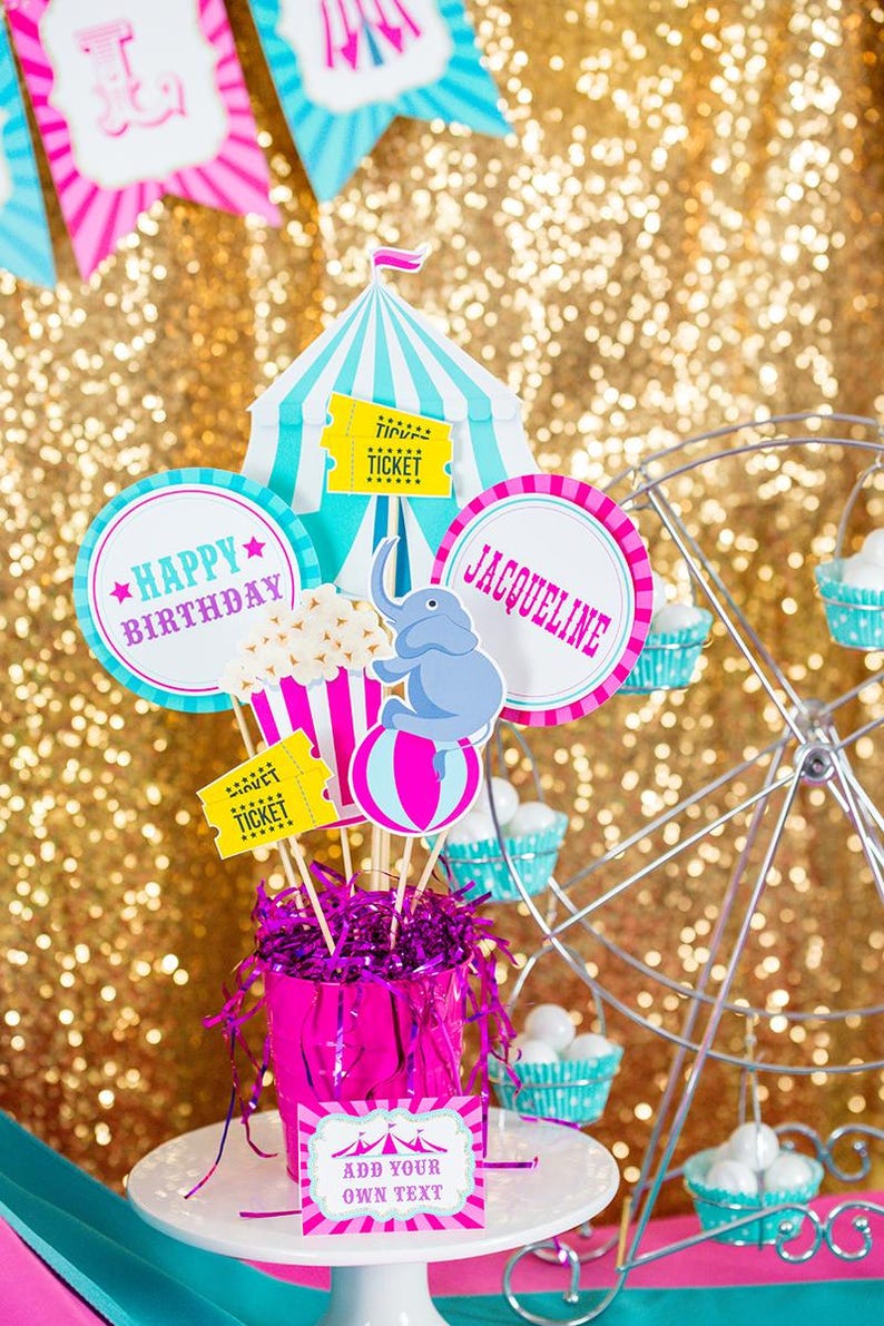 May include: A pink and blue circus-themed birthday party centerpiece with a tent, elephant, popcorn, and tickets. The centerpiece is in a pink bucket with pink and purple tinsel. The text "Happy Birthday" and "Jacqueline" are on the circles. The text "Add your own text" is on a pink and white card.