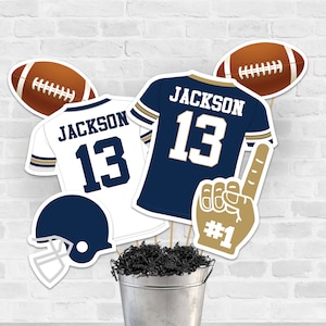 Puede incluir: Un conjunto de seis decoraciones para fiestas con temática de fútbol americano. Las decoraciones incluyen una pelota de fútbol, una camiseta con el nombre "JACKSON" y el número "13", un casco, una camiseta con el nombre "JACKSON" y el número "13", y un dedo de espuma con el número "1".