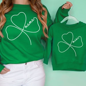 Puede incluir: Camisas de manga larga verdes con un diseño de trébol blanco. La camisa de adulto dice "mama" y la camisa de niño dice "mini".