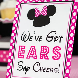 Puede incluir: Un cartel rosa y blanco con lunares y borde negro. El cartel presenta una silueta negra de Minnie Mouse con un lazo rosa de lunares. El texto en el cartel dice "We've Got Ears Say Cheers!"