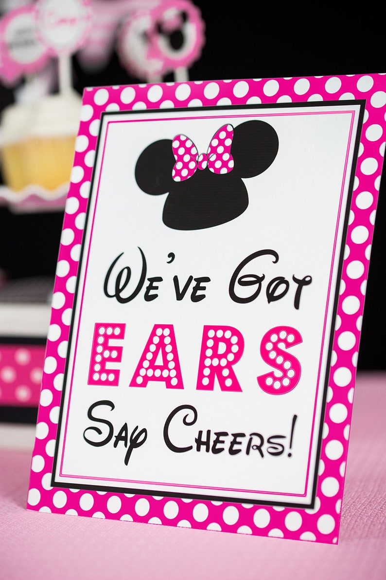 Puede incluir: Un cartel rosa y blanco con lunares y borde negro. El cartel presenta una silueta negra de Minnie Mouse con un lazo rosa de lunares. El texto en el cartel dice "We've Got Ears Say Cheers!"