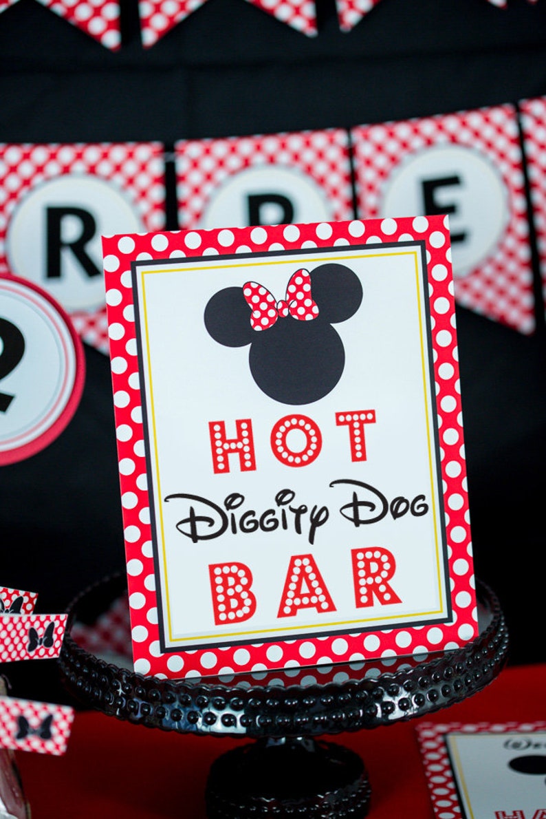以下が含まれることがあります： ミニーマウスのシルエットとリボンが付いた「Hot Diggity Dog Bar」のサイン。サインは赤い水玉模様のボーダーで、黒いケーキスタンドの上にあります。背景には赤と白の水玉模様のバナーがあります。