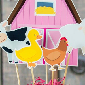 Puede incluir: Un granero rosa con un pajar en la ventana, una vaca blanca, un pato amarillo, una gallina marr&oacute;n y una oveja blanca. Todos los animales est&aacute;n en palos y son parte de un centro de mesa con tem&aacute;tica de granja.