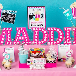 May include: A pink and white polka dot table setting with the name Maddie in large pink letters. The table is decorated with movie-themed party supplies, including popcorn buckets, film reels, and tickets. There are also cupcakes, water bottles, and candy.