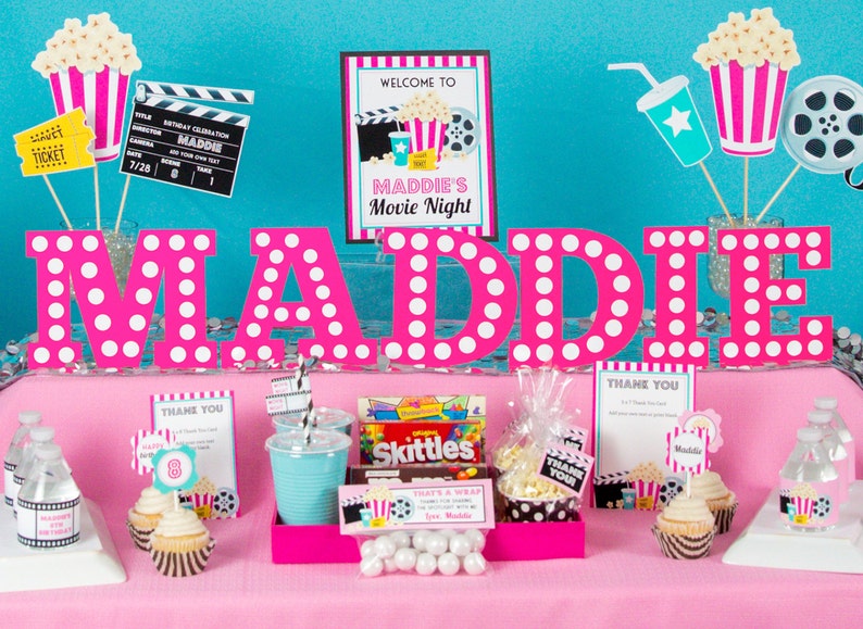 May include: A pink and white polka dot table setting with the name Maddie in large pink letters. The table is decorated with movie-themed party supplies, including popcorn buckets, film reels, and tickets. There are also cupcakes, water bottles, and candy.