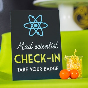 May include: Black sign with a blue atom symbol, the text "Mad scientist check-in" in yellow, and the text "Take your badge" in white.