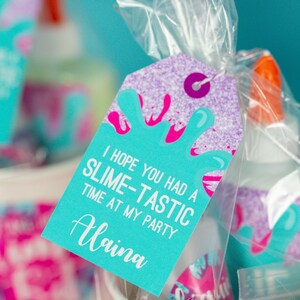 May include: A turquoise blue favor tag with white text that reads "I hope you had a slime-tastic time at my party" and the name "Alaina" in white cursive. The tag has a purple glitter background and a pink and white slime design.