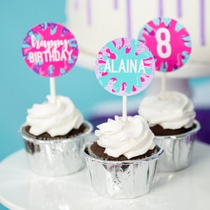 May include: Three cupcakes with white frosting and silver cupcake liners. The cupcakes are decorated with colorful round toppers that say "Happy Birthday", "Alaina", and "8".