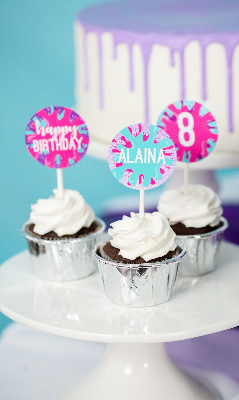 May include: Three cupcakes with white frosting and silver cupcake liners. The cupcakes are decorated with colorful round toppers that say "Happy Birthday", "Alaina", and "8".