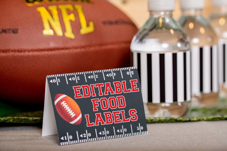 May include: Editable food labels with a football design. The labels are black with red text and a white football graphic. The labels are on a table with a football and water bottles in the background.