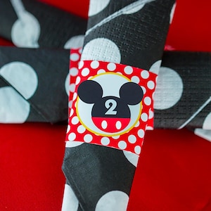 May include: Rolled black napkins with white polka dots, held together by a red band with white polka dots. The band features a Mickey Mouse head and the number 2 in white.