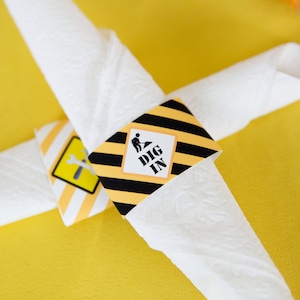 May include: White paper napkin with a black and yellow construction themed napkin ring. The napkin ring has black and yellow stripes and a white square with the text "DIG IN" and a small black silhouette of a construction worker.
