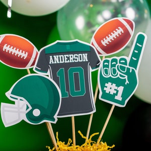 May include: A football-themed centrepiece with a green and white football helmet, a green and white foam finger with the number "#1", a green and white football, a green and white jersey with the name "ANDERSON" and the number "10", and a brown and white football.