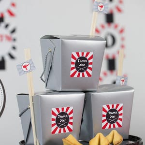 May include: Three silver take-out containers with red and white striped stickers that say "Thank You" with chopsticks and fortune cookies on a black cake stand.