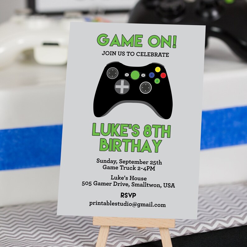 以下が含まれることがあります： 「GAME ON！」と「LUKE'S 8TH BIRTHDAY」の文字が入った灰色の招待状。黒いゲームコントローラーのグラフィックが特徴です。カードには、日付、時間、場所、RSVPのメールアドレスなどのイベント詳細が含まれています。