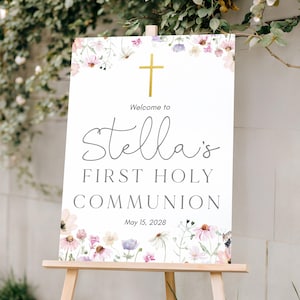 Puede incluir: Un letrero blanco sobre un caballete de madera, decorado con flores de acuarela y una cruz dorada. El letrero dice "Bienvenida a la Primera Comunión de Stella" con la fecha 15 de mayo de 2028.