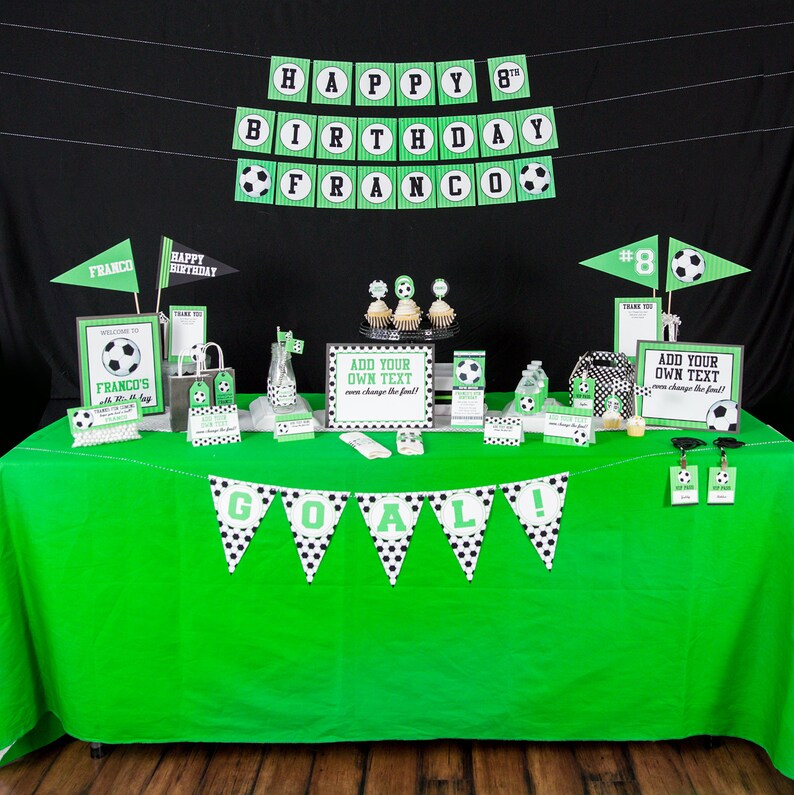 May include: A green and black soccer-themed party table setting with a green tablecloth, black and white polka dot banner that says "GOAL!" and various decorations including a welcome sign, cupcakes, and favor bags.