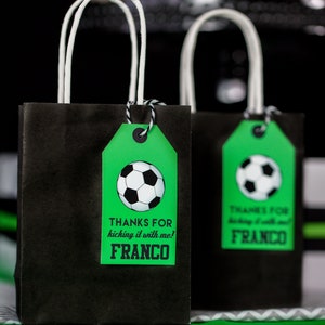Puede incluir: Dos bolsas de regalo negras con asas blancas y etiquetas verdes. Las etiquetas tienen un gr&aacute;fico de un bal&oacute;n de f&uacute;tbol y el texto "Gracias por patear conmigo! Franco".