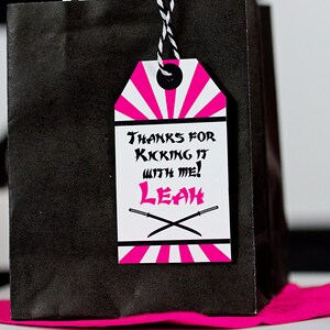 May include: Pink and white favor tag with black text that says "Thanks for kicking it with me! Leah" and a pair of crossed swords.