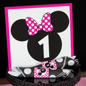 Puede incluir: Un n&uacute;mero de mesa con tem&aacute;tica de Minnie Mouse en rosa y negro con el n&uacute;mero "1" en el centro. El n&uacute;mero de mesa est&aacute; en un soporte de pastel negro con un patr&oacute;n de lunares.