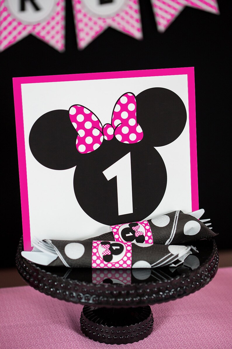 Puede incluir: Un n&uacute;mero de mesa con tem&aacute;tica de Minnie Mouse en rosa y negro con el n&uacute;mero "1" en el centro. El n&uacute;mero de mesa est&aacute; en un soporte de pastel negro con un patr&oacute;n de lunares.