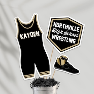 Könnte beinhalten: Schwarz-goldene Partydekorationen zum Thema Wrestling. Enthält einen schwarzen Wrestling-Anzug mit dem Namen "KAYDEN", ein Schild mit der Aufschrift "NORTHVILLE High School WRESTLING" und einen Wrestlingschuh. Die Dekorationen befinden sich in einem silbernen Eimer.
