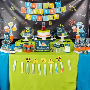 May include: A green tablecloth with a science-themed birthday party setup. The table is decorated with a cake, cupcakes, and various science-themed decorations, including beakers, test tubes, and warning signs. The banner reads "Happy 4th Birthday Waylan".