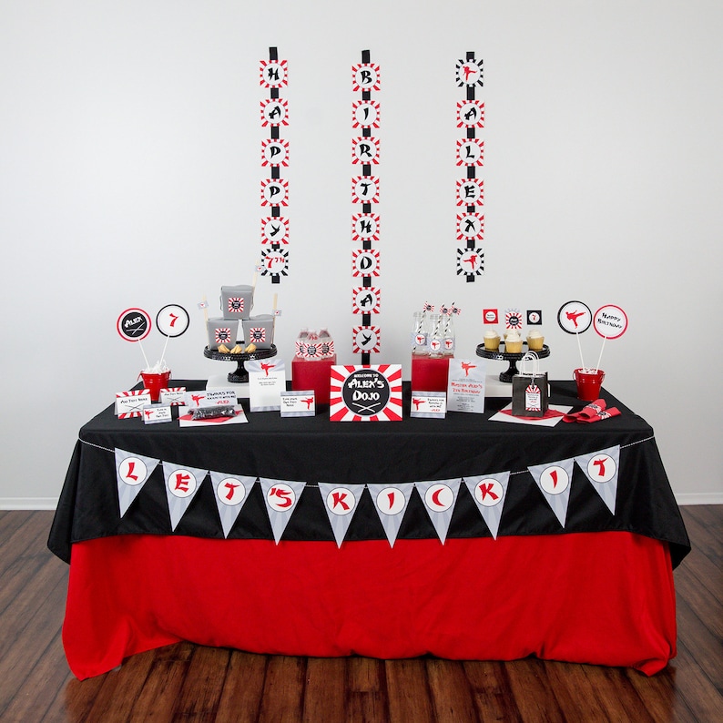 May include: A black and red table with a red tablecloth and a black and white banner that says "Let's Kick It". The table is decorated with a karate-themed birthday party decorations, including a sign that says "Alex's Dojo", black and red paper lanterns, and black and red paper cups.