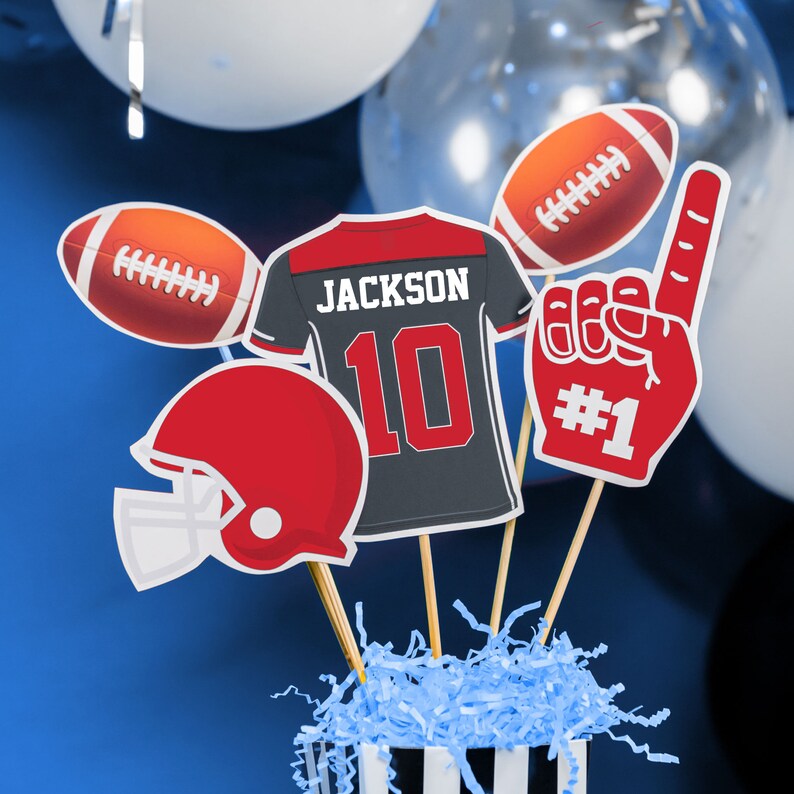Puede incluir: Un centro de mesa con tem&aacute;tica de f&uacute;tbol con una camiseta de f&uacute;tbol roja y negra con el nombre "JACKSON" y el n&uacute;mero "10" en ella, un casco de f&uacute;tbol rojo, un bal&oacute;n de f&uacute;tbol y un dedo de espuma rojo con el n&uacute;mero "#1" en &eacute;l. Los art&iacute;culos est&aacute;n dispuestos en un contenedor a rayas azul y blanco con papel triturado azul.