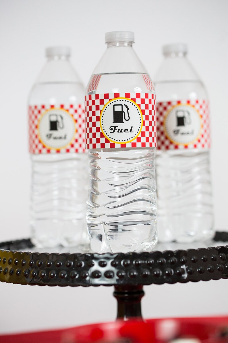 May include: Three clear plastic water bottles with red and white checkered labels that say "Fuel" with a gas pump icon. The bottles are on a black and red tiered stand.