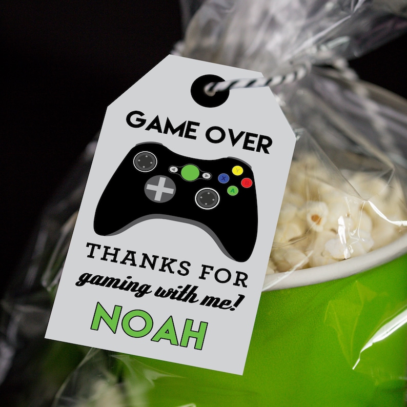 May include: A white favor tag with black and green text. The tag features a black video game controller with green, red, and yellow buttons. The text reads "GAME OVER" and "THANKS FOR gaming with me! NOAH".