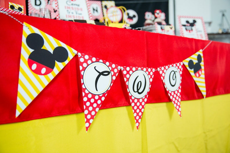 May include: A festive banner with Mickey Mouse-themed pennants. The pennants feature a mix of red polka dots, yellow and white stripes, and Mickey Mouse's iconic silhouette. The banner spells out "TWO" in black script on white circles.
