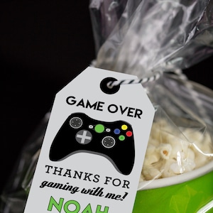 May include: A white favor tag with black and green text that reads "GAME OVER" and features a black video game controller with green, red, yellow, and blue buttons. The tag also says "THANKS FOR GAMING WITH ME! NOAH".