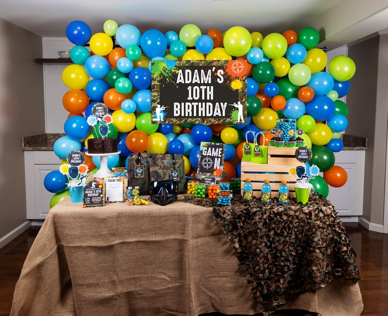 May include: A birthday party table setting with a green, blue, and yellow balloon backdrop. The table is covered in burlap and has a camouflage runner. There are various party decorations, including a cake, candy, and a sign that says "Adam's 10th Birthday".