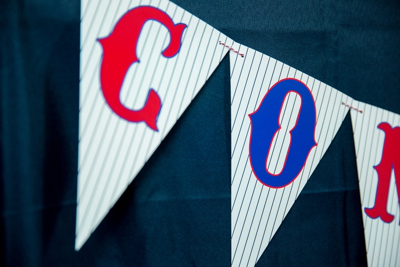 May include: A pennant banner with white and gray pinstripes. The banner features red and blue letters spelling out a word. The letters have a decorative, curved design. The banner is strung with red thread.